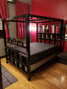 Black metal-framed bondage bed with a stockade at the foot and leather sheets.